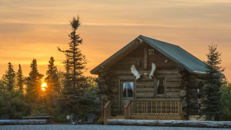 Log cabin with moose antlers above door sits by gravel lot backed by trees in golden sunset at Alaska's Gold Creek Lodge.