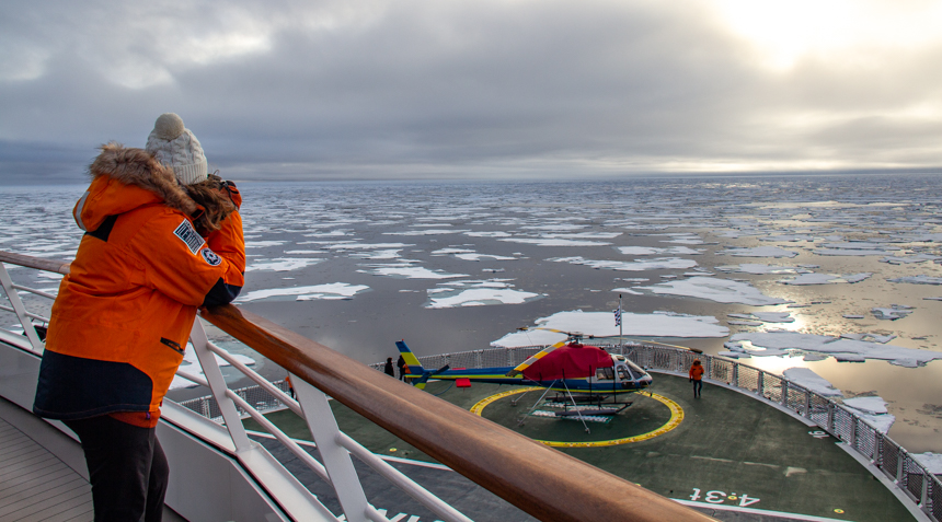 guest in orange parka leans over bow railing using binoculars to look at the endless sea of floating ice sheets. 