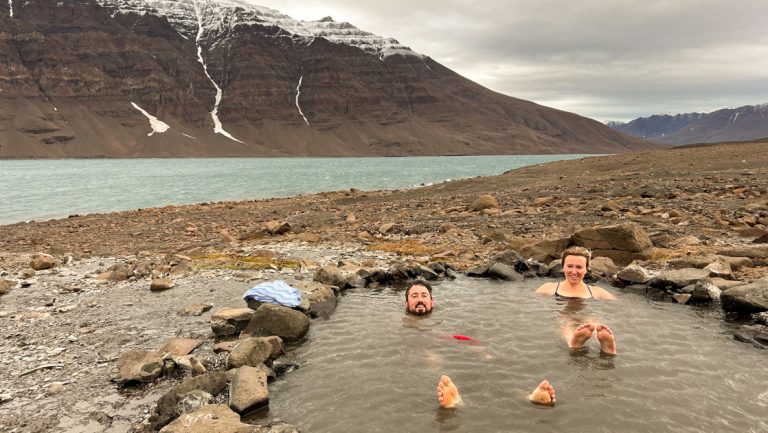 Man & woman sit in natural thermal bath on remote, rocky shoreline under clouds during a cruise to the Faroe Islands & Iceland.