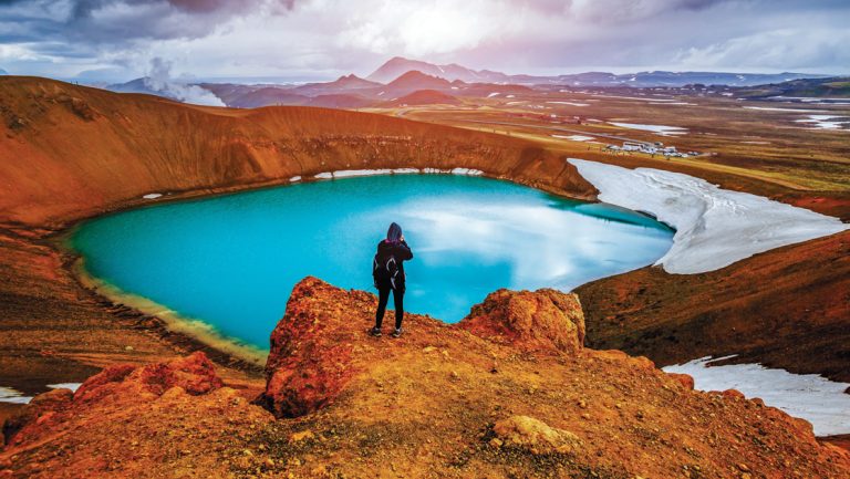 Hiker overlooks geothermal valley with turquoise glacier melt lake & golden-orange hills under cloudy sky by Myvatn Lake, Iceland.