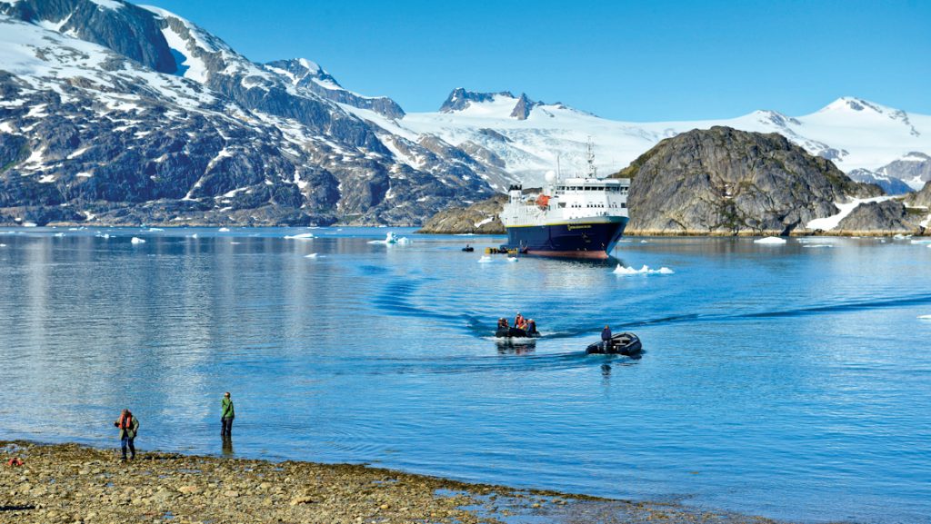 Greenland travelers coming ashore by Zodiac from a small white & blue expedition ship with snowy mountains behind in the sun.