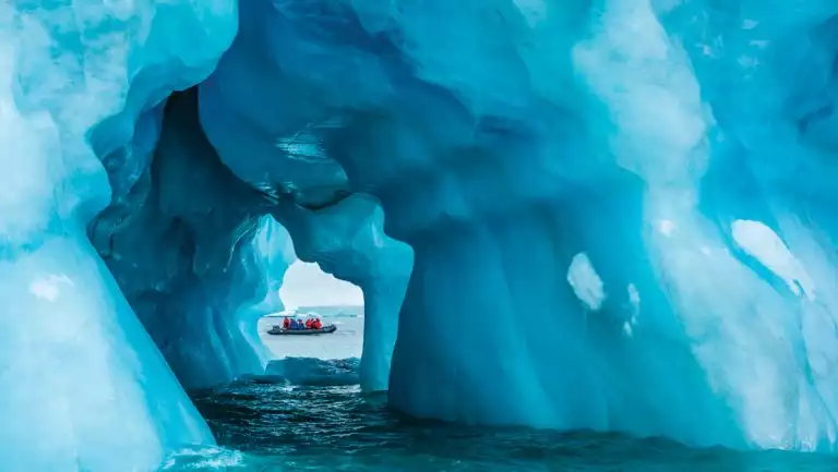 Zodiac dinghy with High Arctic Archipelago cruise guests is viewed through a tunnel in a massive blue iceberg in Greenland.