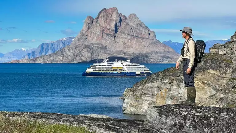 Small Nat Geo ship sits offshore from where man in khaki expedition clothes stands atop beige rocks by green tundra in Greenland.