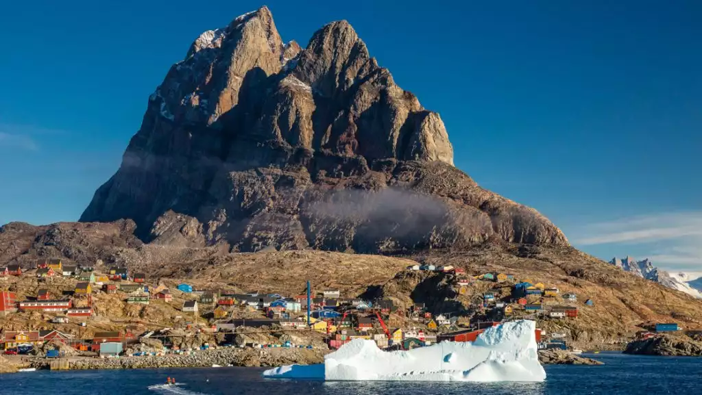 Zodiac boat cruises past big white iceberg towards colorful Arctic town sitting under toothy, rocky mountain in Greenland.