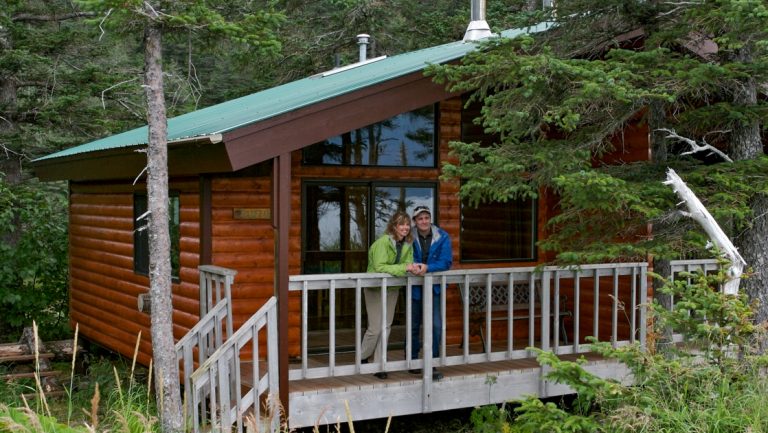 Man & woman stand on deck of guest log cabin with green metal roof at Kenai Fjords Wilderness Lodge on Fox Island, Alaska.