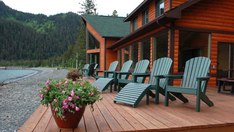 Kenai Fjords Wilderness Lodge's wooden deck with green Adirondack chairs by gray pebble beach, calm sea & green mountains.