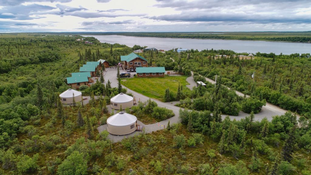 Aerial view of Alaska's Gold Creek Lodge with log cabins with green metal roofs, 3 white yurts, forest & riverside views.