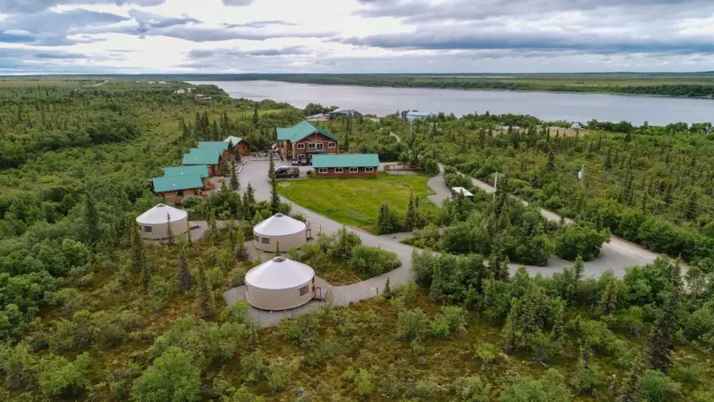 Aerial view of Alaska's Gold Creek Lodge with log cabins with green metal roofs, 3 white yurts, forest & riverside views.