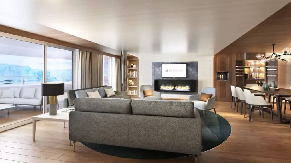 Interior view of the owners suite of a luxury Antarctica cruise ship seen with a couch facing a fireplace, private balcony and dining room with chairs in open concept room