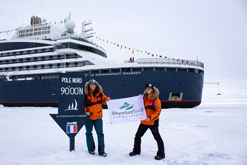Two women in orange parkas stand at north pole sign holding flag in front of white and blue ice breaker ship.