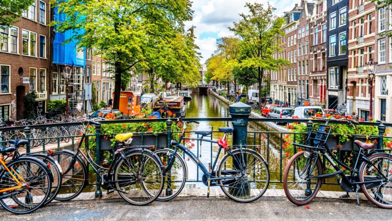 Various commuter bikes parked on bridge overlooking slim canal lined by green trees & colorful apartment buildings in Amsterdam.