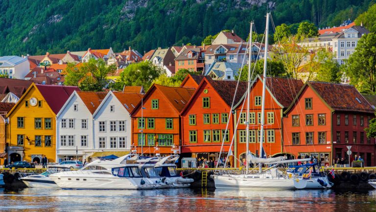 White yacht & sailboat sit in front of row of side-by-side orange, yellow & white houses backed by green hillside in Norway.