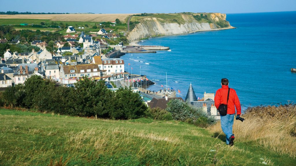 Man in red jacket walks grassy bluff high above the ocean, overlooking a Normandy, France medieval city on North Sea Cruises.
