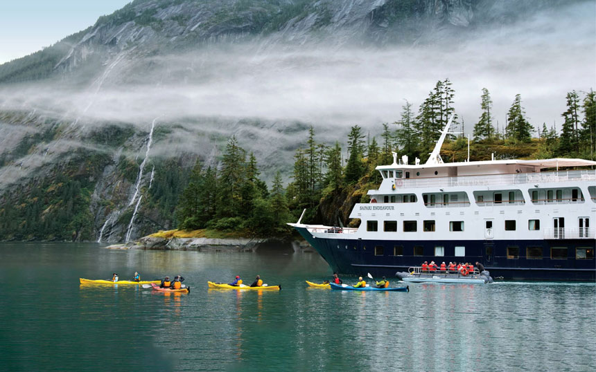 In front of tall fjord walls with waterfalls the white and blue Safari Endeavour ship floats as a group of kayakers paddle yellow and blue kayaks.