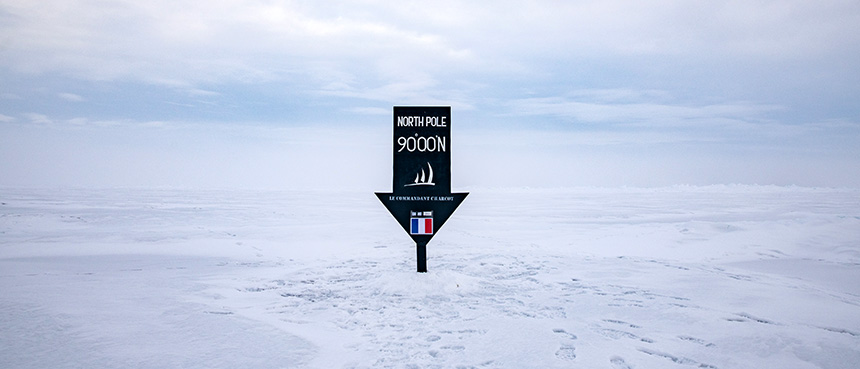 Dark blue down arrow sign with white letters that read North Pole 90.00N planted on ice sheet 