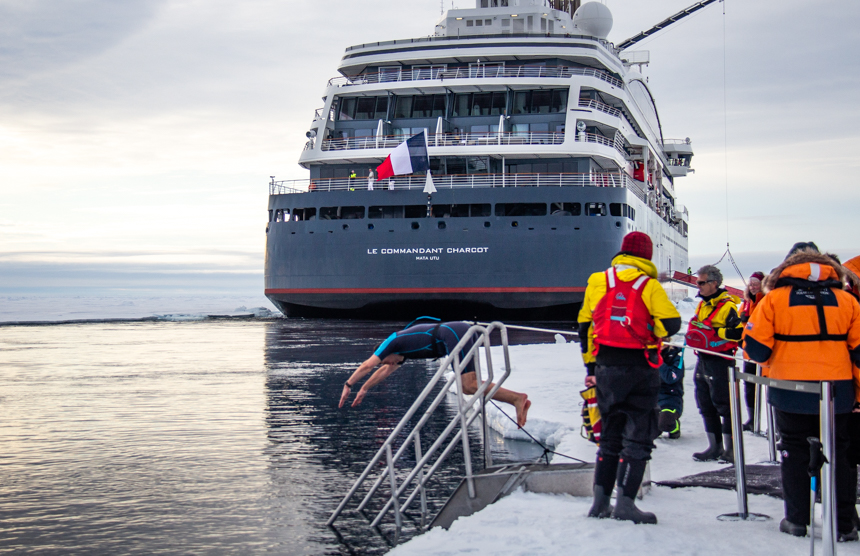 Man in wetsuit dives into ocean from ice edge for polar plunge as Icebreaker ship floats beyond. 