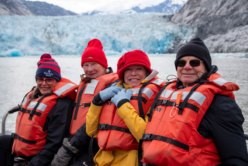 4 Alaska travelers in orange life jackets smile in front of a glacier on a skiff ride on a Prince William Sound cruise.
