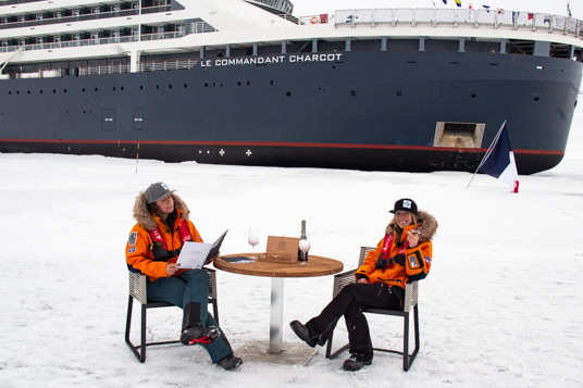 two women in orange parkas sit at the north pole drinking champagne in front of blue and white ice breaker ship