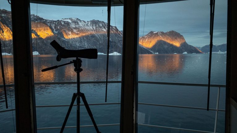 Spotting scope on a tripod sits behind a wall of glass panes on the bridge of a small ship, looking at snowy mountains at dusk.