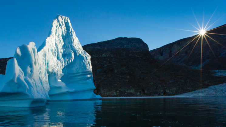Sun bursts above rocky horizon at dawn, lighting a tall iceberg during the Svalbard, Iceland & Greenland's East Coast cruise.