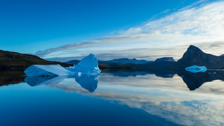 Small icebergs float in glassy water under a partly cloudy sky on a Greenland & Iceland cruise, creating a mirrored reflection.