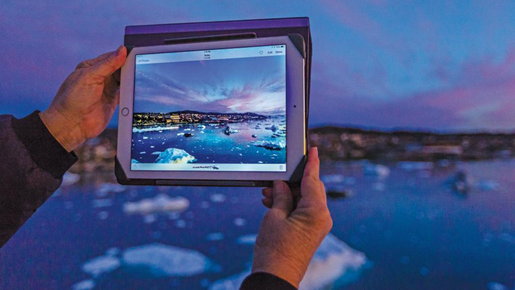 Svalbard, Iceland & Greenland's East Coast traveler holds iPad out to photograph icebergs under a blue & purple sunset.