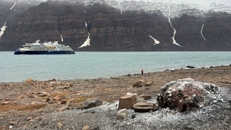 Small cruise ship with dark blue hull & white upper decks sits offshore of a rocky, Arctic beach as solo traveler beachcombs.