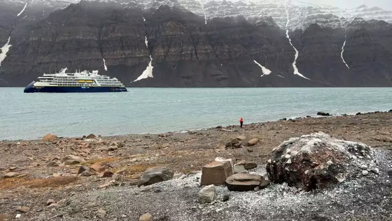 Small cruise ship with dark blue hull & white upper decks sits offshore of a rocky, Arctic beach as solo traveler beachcombs.
