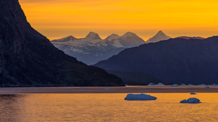 Arctic fjord with jagged peaks behind & small icebergs in the foreground, seen at sunset on a Greenland & Iceland cruise.