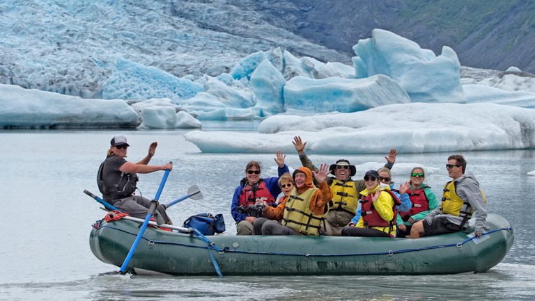 Icy blue raft with travelers & guide floats in calm water past blue glacier & icebergs on the Ultimate Alaska Wildlife Safari.