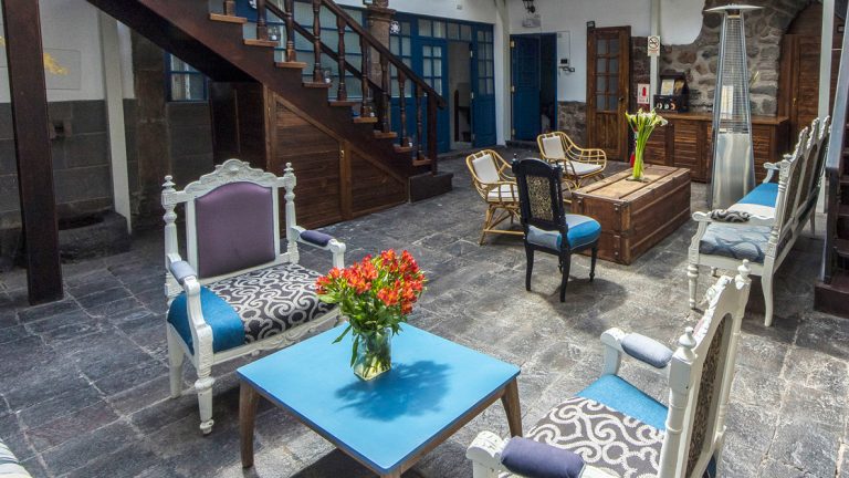 Indoor courtyard of XO Art Hotel Cusco with painted & padded wood chairs around coffee tables on stone floor with natural light.