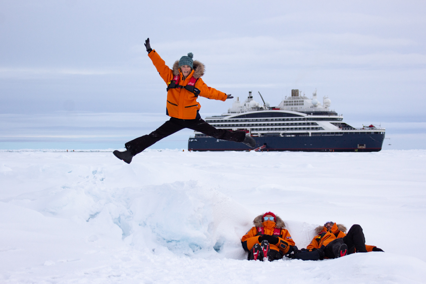 three guests in orange parkas pose and jump in front of ship parked in ice at the North Pole 