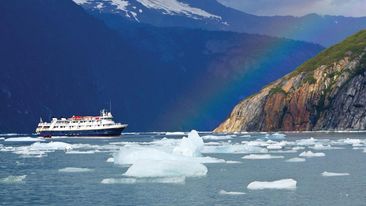 Small ship with dark blue hull & white upper decks sits by iceberg bits in calm water by a rainbow in Prince William Sound.