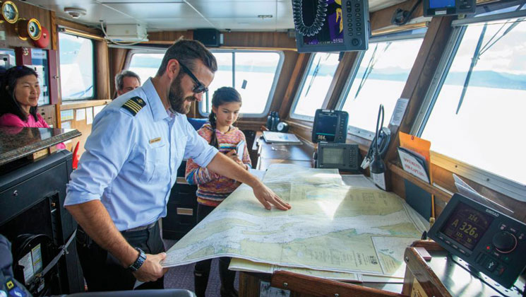 Ship captain explains a nautical map to guests in the bridge of a small ship during a Prince William Sound glacier cruise.