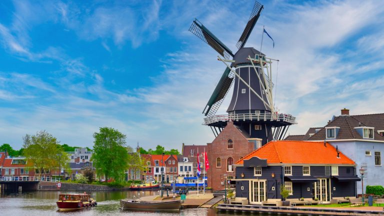 Old-fashioned black windmill sits beside canal among multicolored, short buildings under a blue sky in Amsterdam.