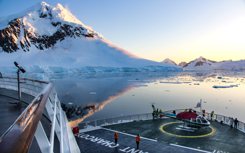 Bow of ship with guests and helicopter, overlooking a pastel colored sunset over snow covered Antarctica landscape.