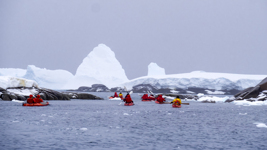 Group of kayakers in red parkas and red double kayaks paddle in the dark waters of Antarctica around icebergs on an overcast day. 