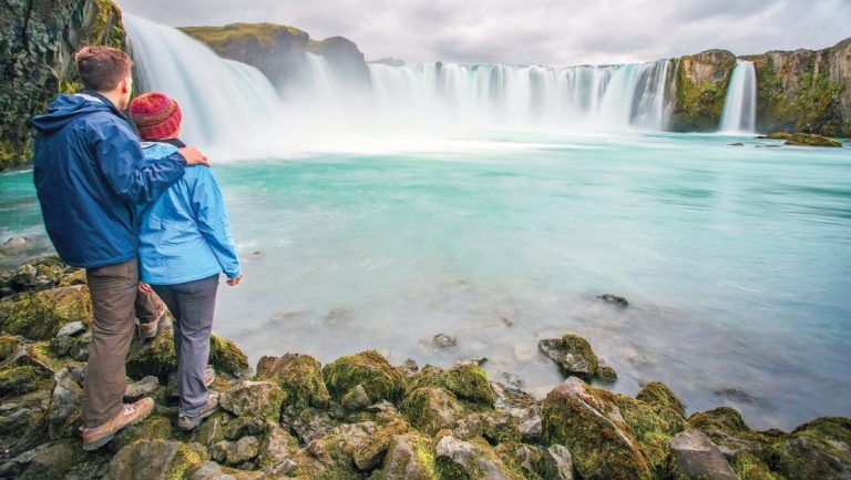 A couple stand together viewing big Godafoss waterfall with teal water in Iceland during a Legendary Northern Isles cruise.