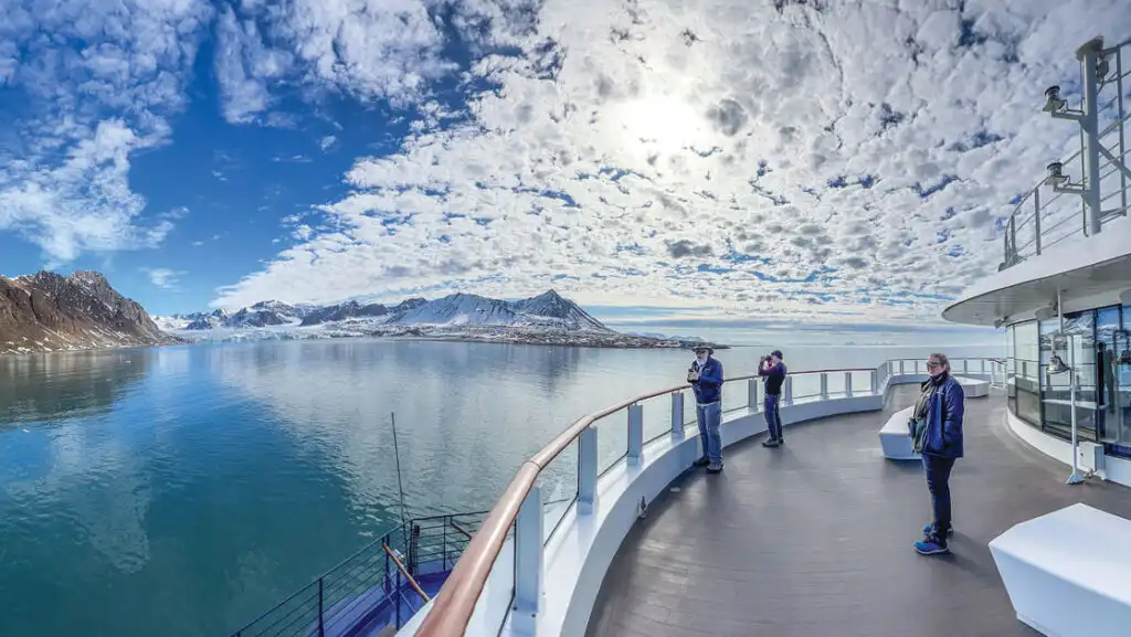 3 travelers stand on deck of a modern ship viewing rocky & snowy peaks & a large white glacier on a Norwegian Discovery cruise.