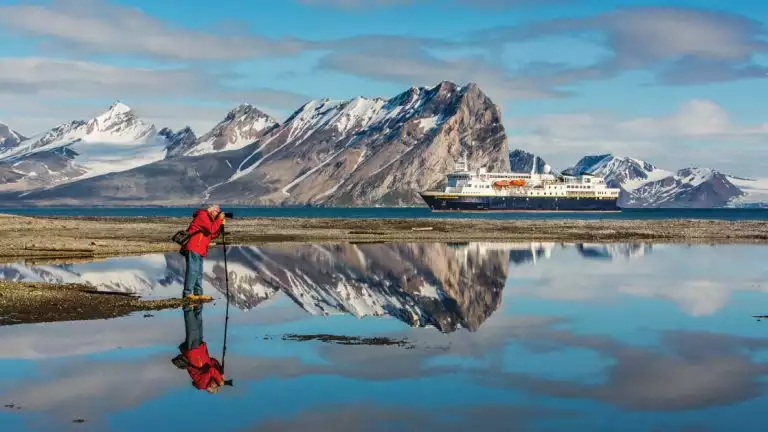 traveler takes a picture from a glassy pond in front of the National Geographic Explorer arctic expedition ship in the background in the Svalbard Archipelago, Norway.