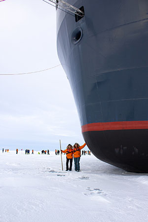 Two travelers at the North Pole stand beneath the bow of their expedition ship as it's parked on the sea ice