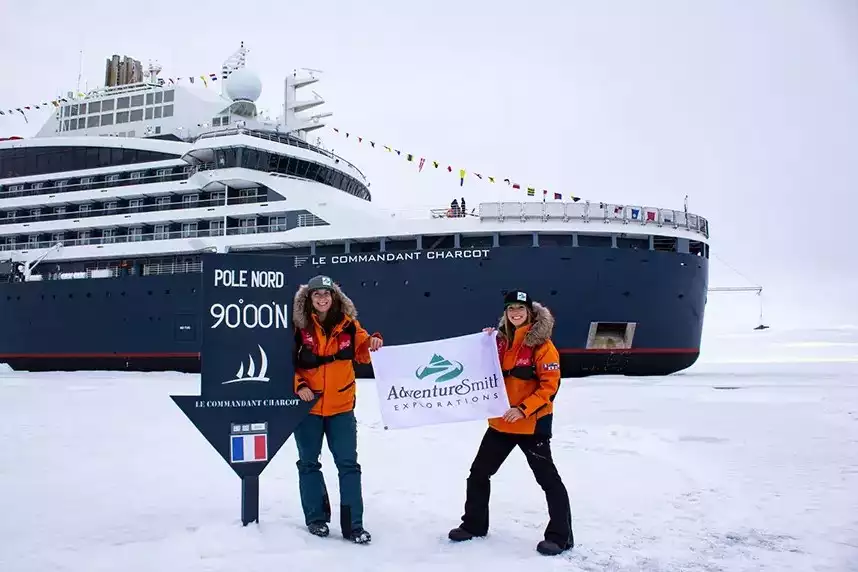Two travelers in orange jackets stand at the North Pole 90 degrees blue sign in front of an expedition ship and holding an AdventureSmith Explorations flag between them