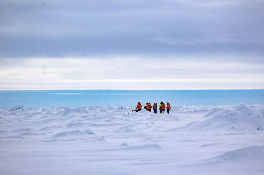 A small group of North Pole cruise passengers stands on sea ice amid mini peaks of snow and ice with the blue ocean behind them