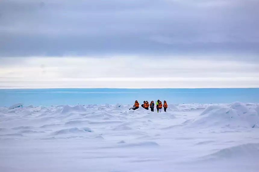A small group of North Pole cruise passengers stands on sea ice amid mini peaks of snow and ice with the blue ocean behind them