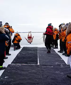A man does a flip into the icy waters at the North Pole with other travelers watching him and black mats covering the ice