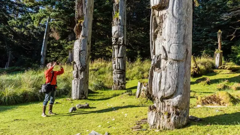Man in red jacket stands in grass & photographs weathered totem poles on an Alaska, BC & Canada small ship cruise.