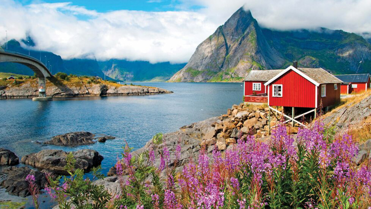 Bright red house on stilts sits atop rocky shoreline with tall pink flowers & fjords in the background on a partly sunny day.