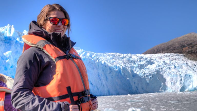 Woman in red sunglasses & orange life jacket stands in a Zodiac boat beside a large white & blue iceberg in Southern Patagonia.