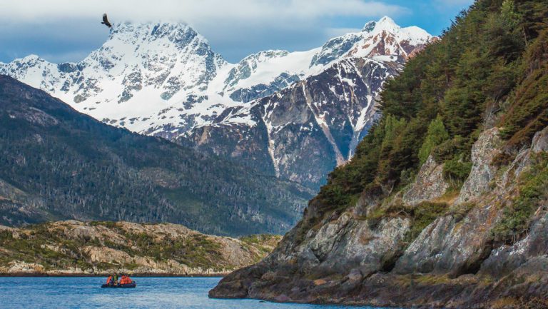 Zodiac of Southern Patagonia cruise travelers motors through a large channel among towering mountains with green vegetation.