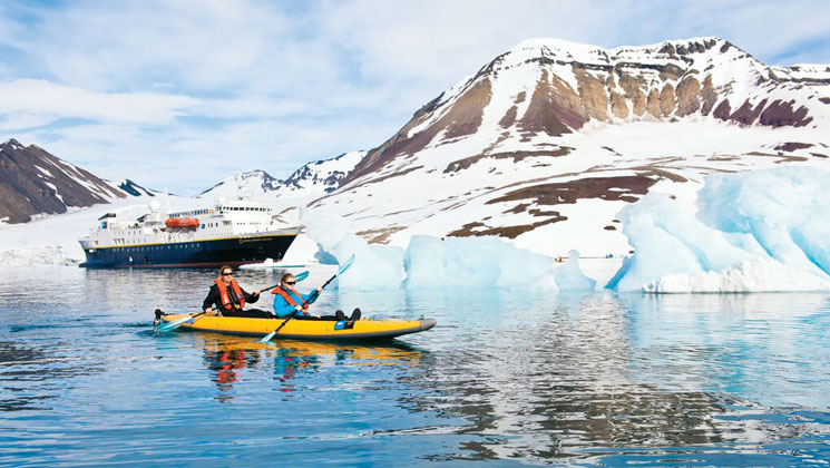 Woman & child paddle tandem yellow inflatable kayak in glassy water beside small icebergs & snow-covered brown mountains.
